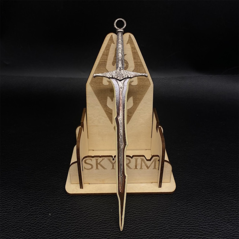 Puede incluir: Una espada detallada de color plateado con empu&ntilde;adura y hoja decorativas, expuesta sobre un soporte de madera. El soporte presenta la palabra "SKYRIM" y un logotipo. La espada tiene un anillo en la parte superior.