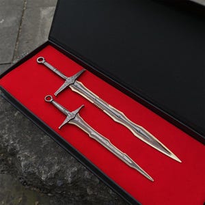 May include: Two decorative metal swords with ornate handles and blades, presented in a black box with a red velvet lining. The swords have a silver and bronze color scheme, with detailed crossguards and pointed tips. The box is open, showcasing the swords.