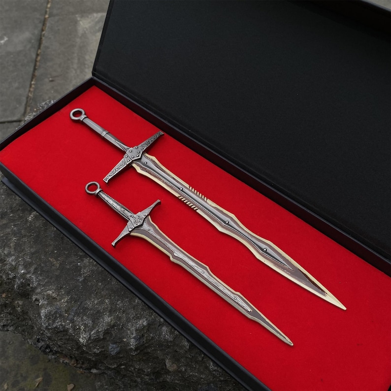 May include: Two decorative metal swords with ornate handles and blades, presented in a black box with a red velvet lining. The swords have a silver and bronze color scheme, with detailed crossguards and pointed tips. The box is open, showcasing the swords.