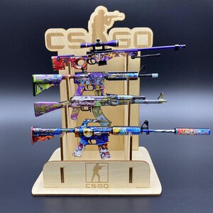 May include: A wooden display stand holding four toy rifles with colorful, graffiti-style designs. The rifles are in shades of purple, blue, and green, with intricate patterns. The stand has the text "CS:GO" and a silhouette of a person.