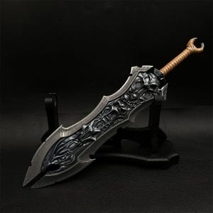 May include: A detailed, fantasy-style sword with a dark, metallic finish. The blade features intricate carvings and a skull motif. The handle is wrapped in a tan material, and the sword rests on a black display stand.