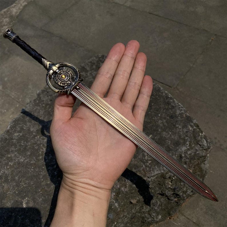 May include: A decorative sword with a bronze-colored hilt and a blade with gold and red accents. The hilt features intricate circular designs and a dark brown, textured handle. The sword is held in a hand.