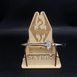 May include: A wooden display stand with the word "SKYRIM" engraved on the front. The stand holds a decorative sword with a silver blade and a brown handle. The stand features a dragon emblem.