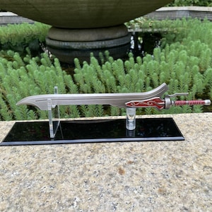 Könnte beinhalten: Ein dekoratives Schwert mit einer silbernen Klinge und einem roten Griff. Das Schwert wird auf einem transparenten Ständer mit schwarzem Sockel ausgestellt. Der Griff weist aufwendige Details auf, und die Klinge hat ein gebogenes Design.