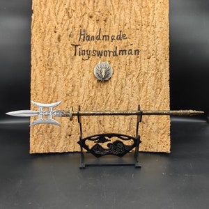 Op de afbeelding: Een decoratieve speer met een gouden handvat en een zilveren blad. De speer is gemonteerd op een zwarte standaard met een golfontwerp. De standaard staat op een kurk achtergrond met de tekst "Handmade Tinyswordman".