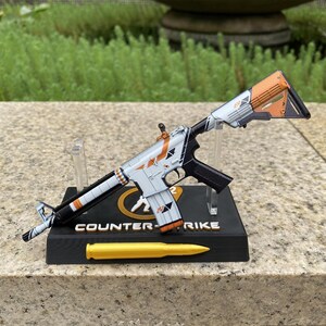 May include: A miniature rifle model featuring a white, black, and orange colour scheme, mounted on a black display stand labelled Counter-Strike. The base includes a gold-coloured bullet replica, and the rifle body displays geometric patterns.