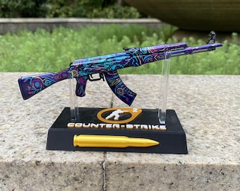 May include: A miniature rifle model featuring a vibrant purple, blue, and pink swirl pattern. It is mounted on a black display stand with the text "COUNTER-STRIKE" and a "2" logo, accompanied by a single gold-colored bullet replica.