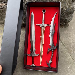 May include: A set of three miniature decorative swords in a black box with a red interior. The swords have silver and bronze tones, with intricate handle and blade designs. The box is open, revealing the swords.