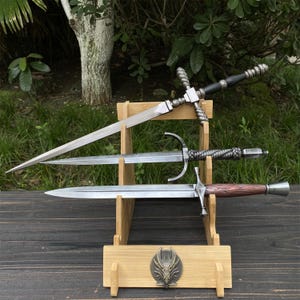 Può includere: Tre spade esposte su un supporto di legno. La spada superiore ha un'impugnatura nera e una guardia trasversale. La spada centrale ha una lama curva e un'impugnatura intrecciata. La spada inferiore ha un'impugnatura marrone.