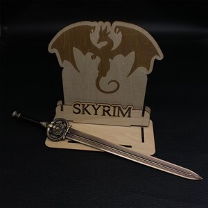 May include: A wooden display stand with a dragon silhouette and the word "SKYRIM" laser-cut into it. A bronze-colored sword with intricate detailing rests in front of the stand, showcasing a fantasy theme.