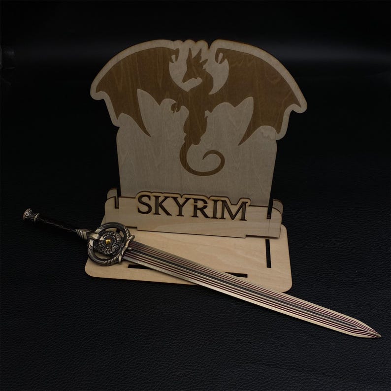 May include: A wooden display stand with a dragon silhouette and the word "SKYRIM" laser-cut into it. A bronze-colored sword with intricate detailing rests in front of the stand, showcasing a fantasy theme.