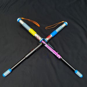 May include: Four colorful, extendable walking sticks are crossed against a black background. Each stick has a different color scheme, including blue, yellow, pink, and silver accents. They have wrist straps and are designed for outdoor activities.