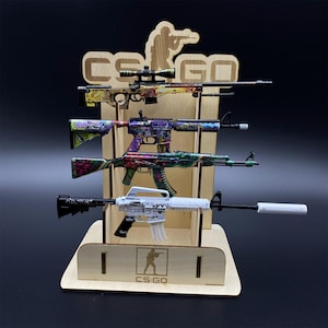 Puede incluir: Un expositor de madera con cuatro armas de juguete con dise&ntilde;os coloridos. El expositor tiene el texto "CS:GO" y una silueta de una persona sosteniendo un arma. Las armas de juguete presentan diversos patrones y colores.