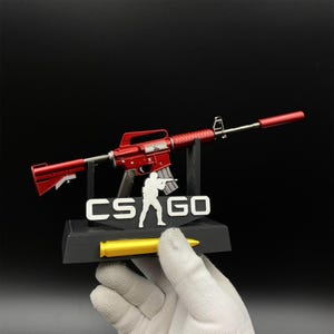 Pu&ograve; includere: Un fucile giocattolo rosso con silenziatore, esposto su un supporto nero con la scritta "CS GO" e una silhouette bianca di una persona che tiene in mano una pistola. Un proiettile giallo &egrave; di fronte al supporto.