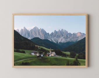 May include: A framed landscape photograph featuring a small village with a church nestled in a lush green valley, set against a backdrop of jagged, towering grey mountain peaks under a clear, pale blue sky during the soft light of day.