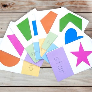 May include: A set of colorful flashcards featuring different shapes, including a circle, square, rectangle, triangle, heart, star, and arrow. Each card has the name of the shape written on it.
