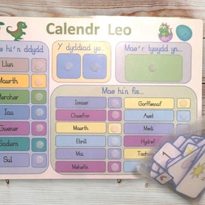 May include: A colorful children's calendar with dinosaur illustrations and Welsh text. The calendar includes sections for the day, date, and weather, with a set of weather cards in a small bag.