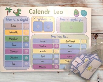 May include: A colorful children's calendar with dinosaur illustrations and Welsh text. The calendar includes sections for the day, date, and weather, with a set of weather cards in a small bag.