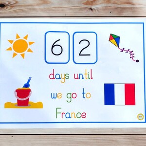 May include: A white countdown board with a blue border shows 62 days until we go to France. The board has a yellow sun, a red bucket and shovel, a blue and red kite, and a French flag.