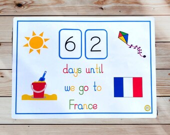 May include: A white countdown board with a blue border shows 62 days until we go to France. The board has a yellow sun, a red bucket and shovel, a blue and red kite, and a French flag.