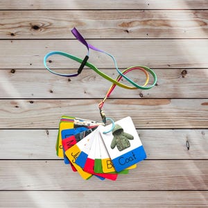 May include: A set of colorful flashcards with images and words for learning about clothing. The cards are attached to a rainbow lanyard. The cards include words like "Coat", "Boots", "Jumper", "Shirt", and "Hat".