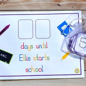 May include: A white countdown board with purple trim, featuring spaces for numbers, a crayon, and a chalkboard. Text reads "days until Ellis starts school." A small bag contains number cards. A yellow pencil graphic is also present.