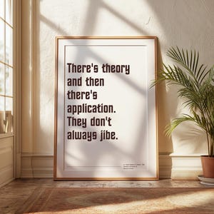 May include: Framed print with a gold border, featuring the quote "There's theory and then there's application. They don't always jibe." The text is in a bold, serif font against a white background. A potted plant sits to the right.