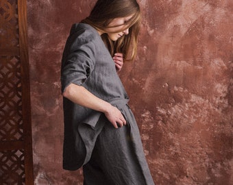 May include: A woman wearing a gray linen pantsuit. The pantsuit has a relaxed fit and is made of a lightweight fabric. The woman is standing in front of a textured wall.