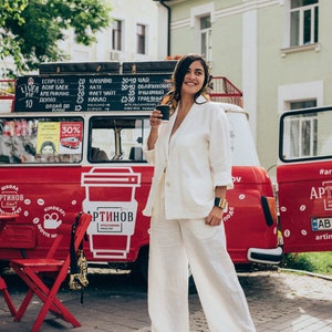 May include: A woman in a white linen suit stands in front of a red food truck with a coffee cup in her hand. The truck has a sign that says "Artinov Creative Space" and a logo of a coffee cup. The woman is smiling and looking at the camera.