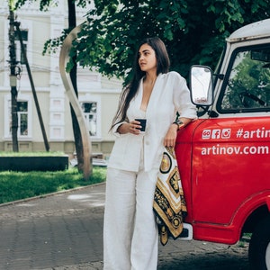 May include: A woman in a white pantsuit stands next to a red vintage van. She is holding a black cup and has a black and gold patterned scarf draped over her arm. The van has the text "go fo #artinov.com" on the side.