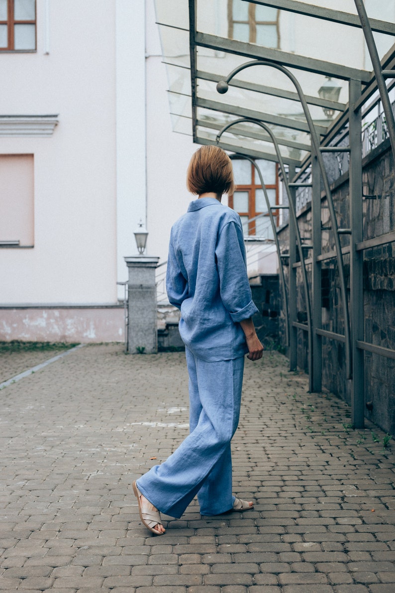 May include: A person wearing a light blue linen jumpsuit walks on a brick pathway. The person is facing away from the camera and has short brown hair.