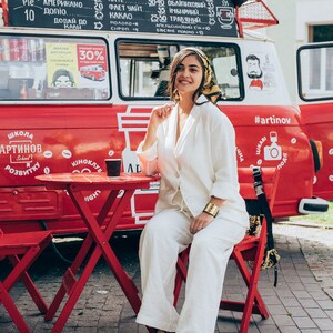 May include: A woman wearing a white pantsuit and a headscarf sits on a red chair in front of a red food truck. The truck has a sign with the words "Школа Артинов" and "#artinov" written on it. The woman is holding a cup of coffee in her hand.