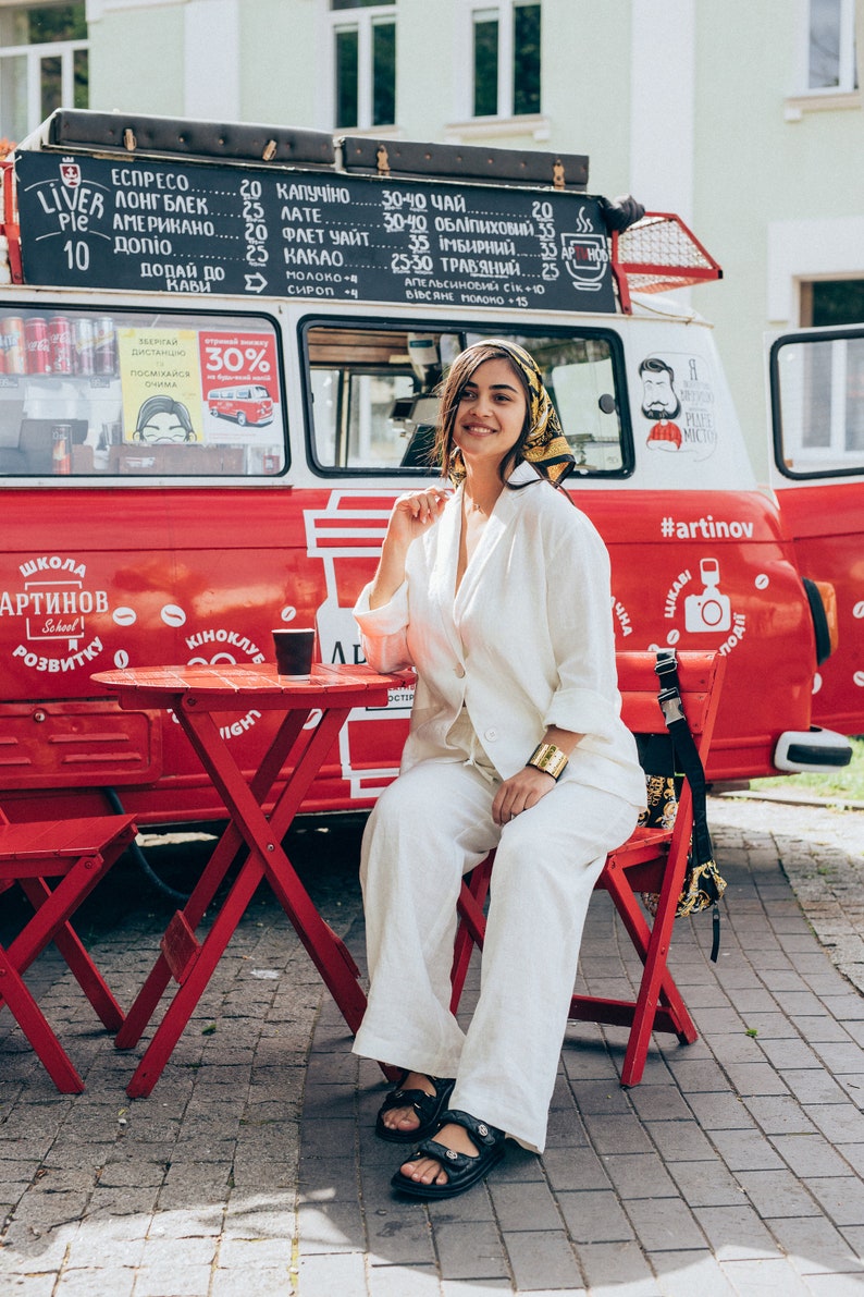 May include: A woman wearing a white pantsuit and a headscarf sits on a red chair in front of a red food truck. The truck has a sign with the words "Школа Артинов" and "#artinov" written on it. The woman is holding a cup of coffee in her hand.