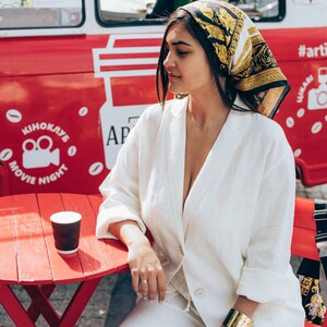 May include: A woman wearing a white linen jumpsuit and a black and gold patterned headscarf sits at a red table with a disposable coffee cup. The table is in front of a red and white food truck with the text "KINOCLUB MOVIE NIGHT" and "ART" on the side.