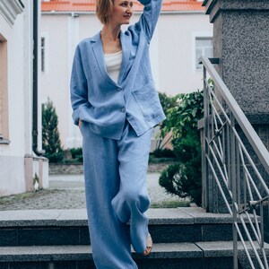 May include: A woman wearing a light blue linen pantsuit standing on a set of stone steps. The pantsuit has a blazer with a single button closure and wide-leg pants.