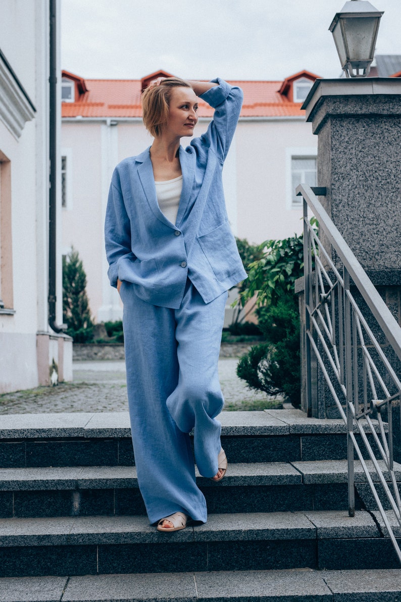 May include: A woman wearing a light blue linen pantsuit standing on a set of stone steps. The pantsuit has a blazer with a single button closure and wide-leg pants.