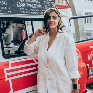 May include: A woman wearing a white linen blazer and pants stands in front of a red food truck with a sign that reads "ARTINO - CREATIVE SPACE". The truck has a menu board with prices for various drinks, including tea, herbal teas, and juices.