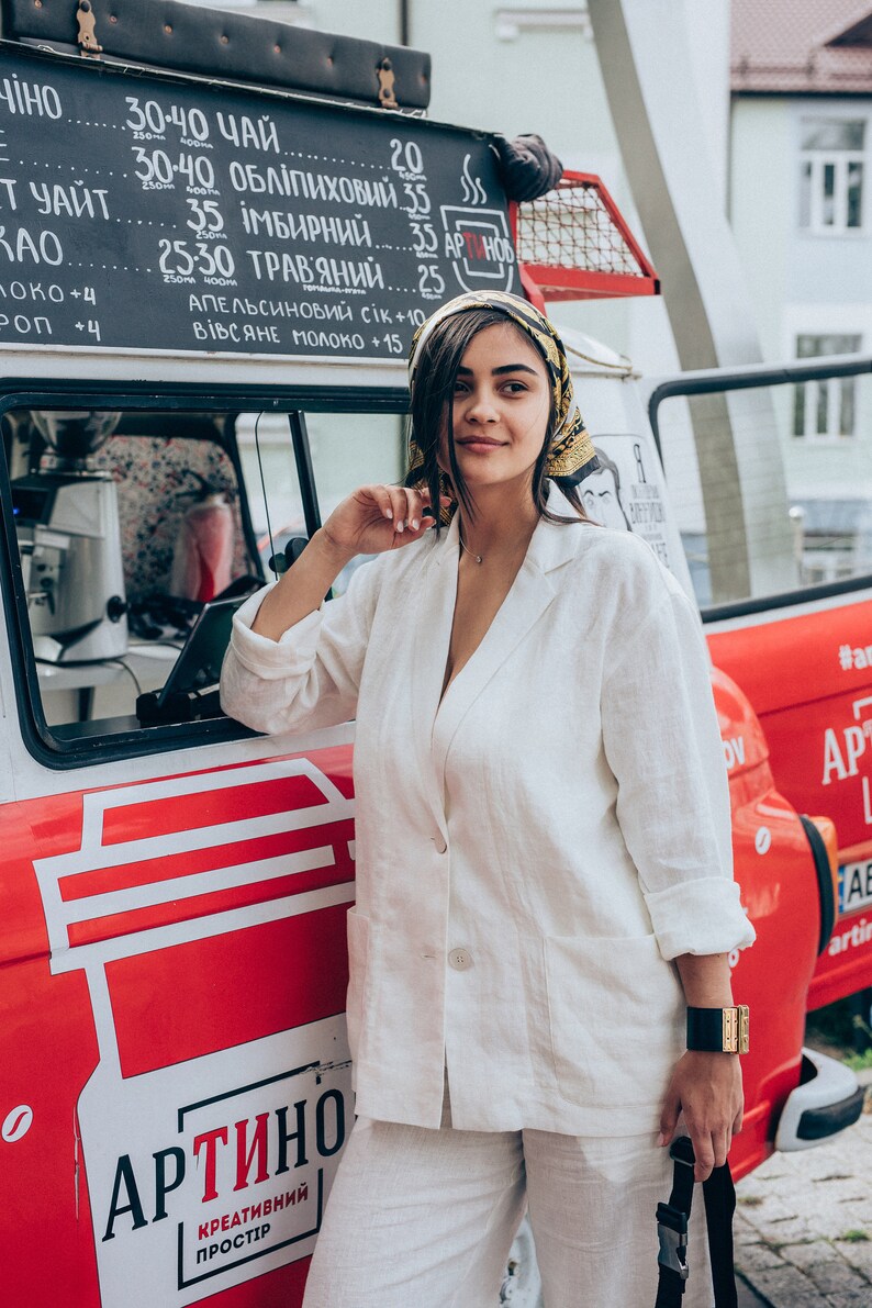 May include: A woman wearing a white linen blazer and pants stands in front of a red food truck with a sign that reads "ARTINO - CREATIVE SPACE". The truck has a menu board with prices for various drinks, including tea, herbal teas, and juices.