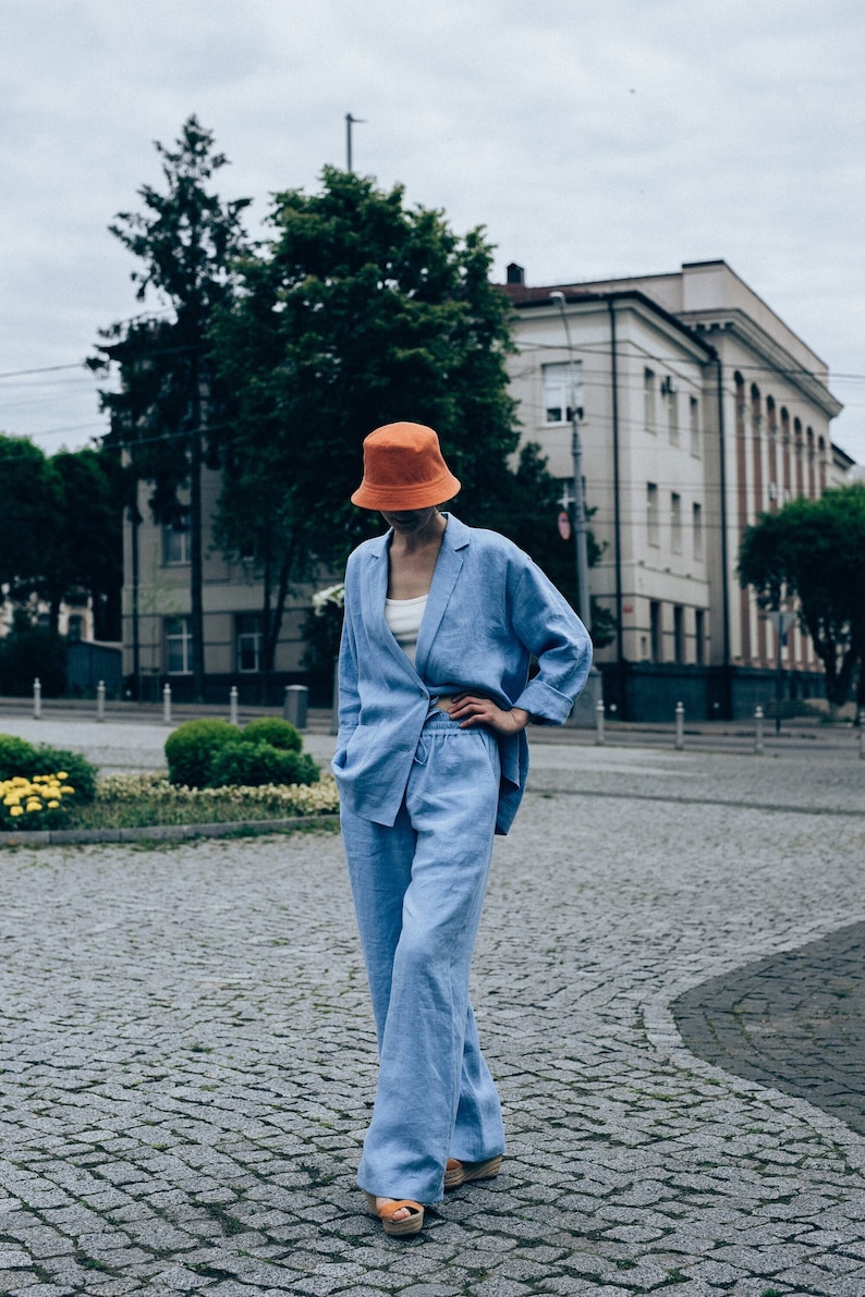 May include: A person wearing a light blue linen pantsuit with a matching blazer and wide-leg pants. They are wearing an orange bucket hat and standing on a cobblestone street.
