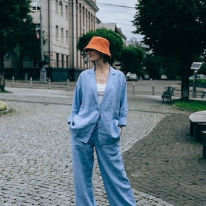 May include: A woman wearing a light blue linen suit with a matching blazer and pants. She is wearing a bright orange bucket hat and brown sandals. The woman is standing on a cobblestone street in front of a building.