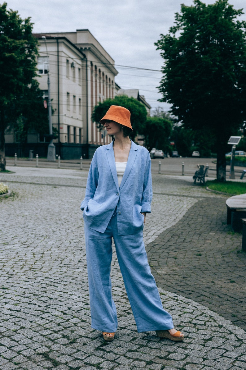May include: A woman wearing a light blue linen suit with a matching blazer and pants. She is wearing a bright orange bucket hat and brown sandals. The woman is standing on a cobblestone street in front of a building.