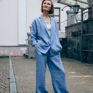 May include: A woman wearing a light blue linen pantsuit. She is standing on a brick walkway and looking up.
