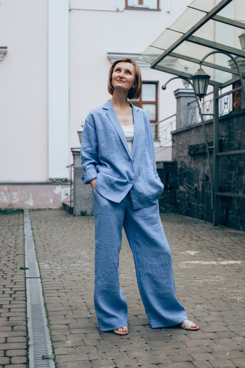 May include: A woman wearing a light blue linen pantsuit. She is standing on a brick walkway and looking up.