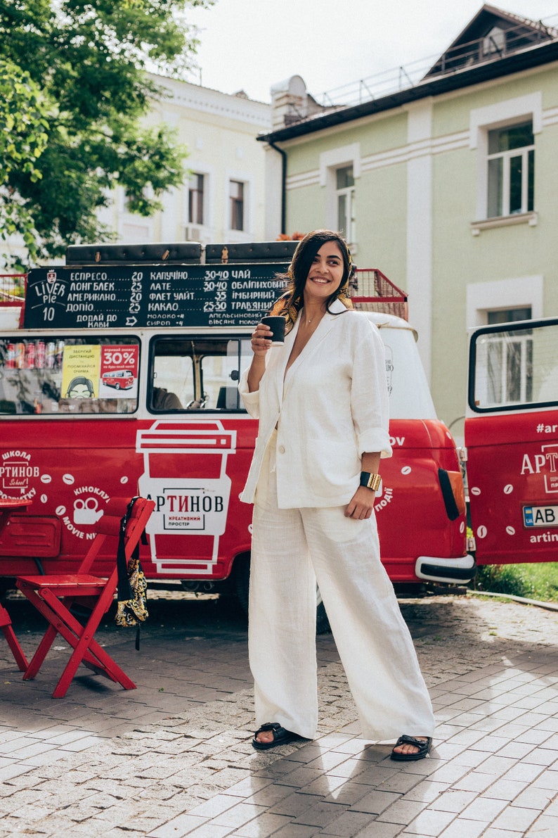 May include: A woman in a white linen suit stands in front of a red food truck with a coffee cup in her hand. The truck has a sign that says "Artinov Creative Space" and a logo of a coffee cup. The woman is smiling and looking at the camera.