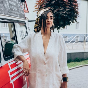 May include: A woman wearing a white linen blazer and matching pants stands next to a red food truck. She is wearing a gold and black patterned headscarf and a gold necklace. The truck has a sign that says "АРТИНОВ" in black lettering.