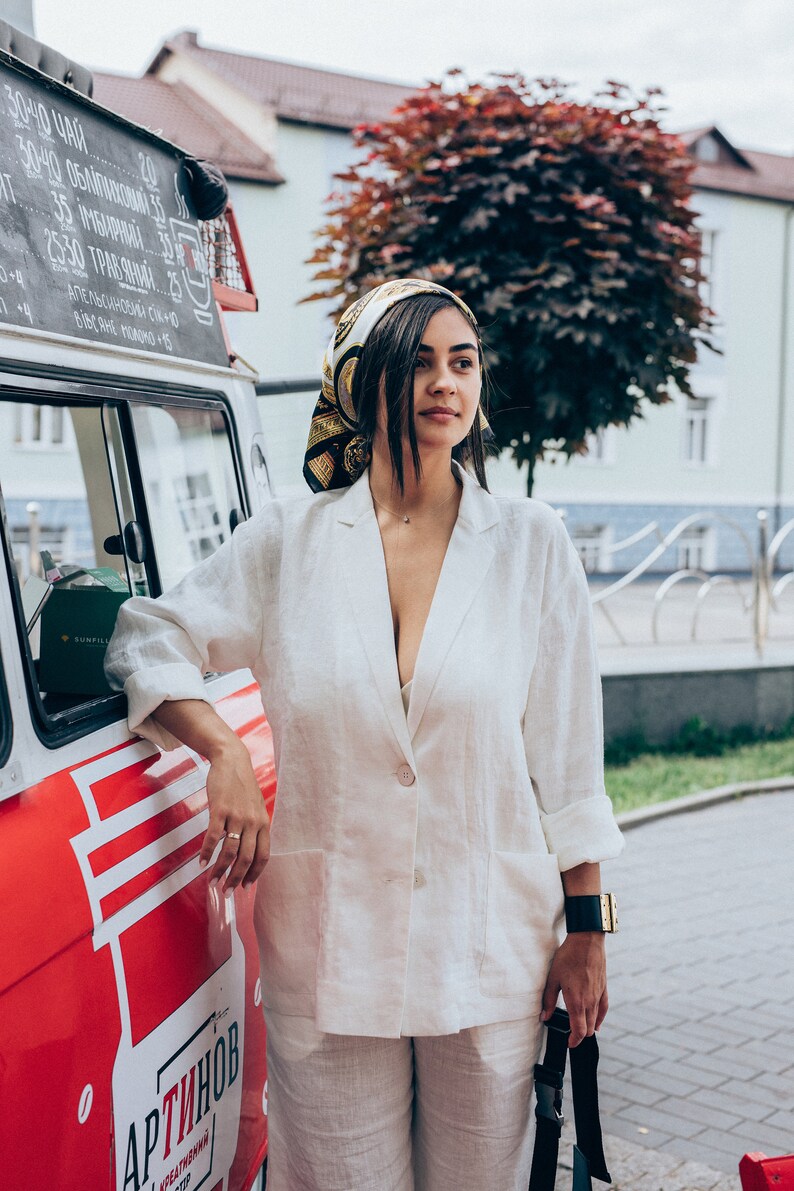May include: A woman wearing a white linen blazer and matching pants stands next to a red food truck. She is wearing a gold and black patterned headscarf and a gold necklace. The truck has a sign that says "АРТИНОВ" in black lettering.