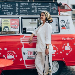 May include: A woman wearing a white linen pantsuit and a patterned headscarf stands in front of a red food truck. The truck has a sign that says "Artino" and "Creative Space" in Ukrainian. The truck is decorated with coffee beans and other designs.