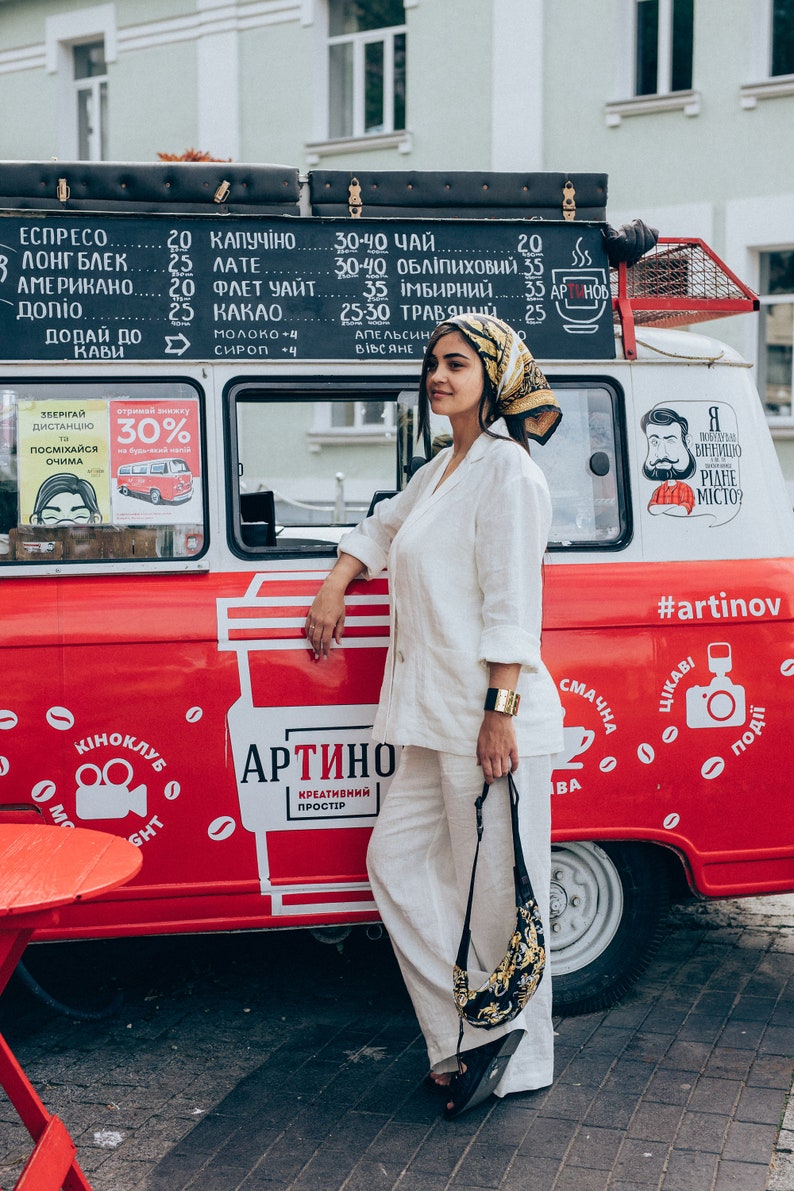 May include: A woman wearing a white linen pantsuit and a patterned headscarf stands in front of a red food truck. The truck has a sign that says "Artino" and "Creative Space" in Ukrainian. The truck is decorated with coffee beans and other designs.
