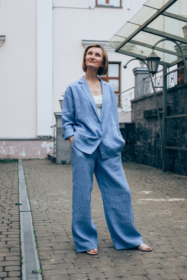 May include: A woman wearing a light blue linen pantsuit. The pantsuit has a blazer with a single button closure and wide-leg pants. The woman is standing on a brick walkway.