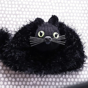 May include: A handmade black cat plush toy with green eyes and whiskers. The cat is made of knitted yarn and fluffy black material. The background features a white and grey polka dot pattern.
